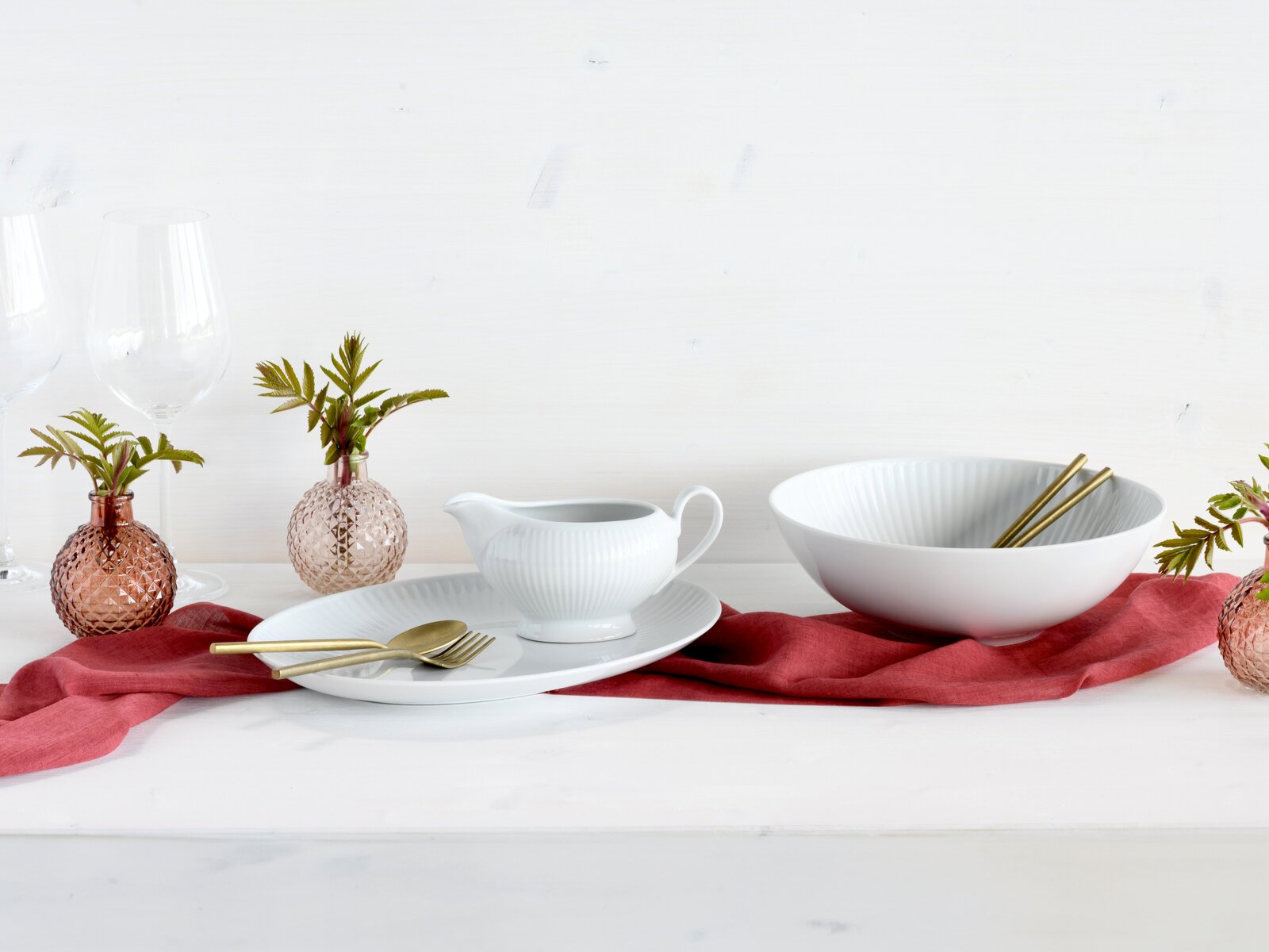 Creatable Servierset 3-tlg VALENCIA Weiße, geriffelte Keramik-Schale mit goldfarbenen Essstäbchen, daneben weißes Soßenschälchen auf Teller mit goldfarbenem Besteck auf rotem Leinentuch; Perspektive: seitliche Frontansicht auf Tischhöhe vor hellem Hintergrund.