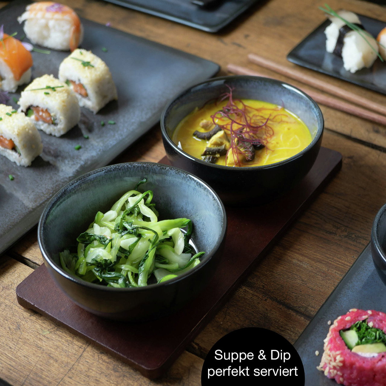 Nahaufnahme eines asiatischen Sushi-Sets mit blauen Tellern und Schalen, serviert mit Suppe und Dip auf einem Holztisch.