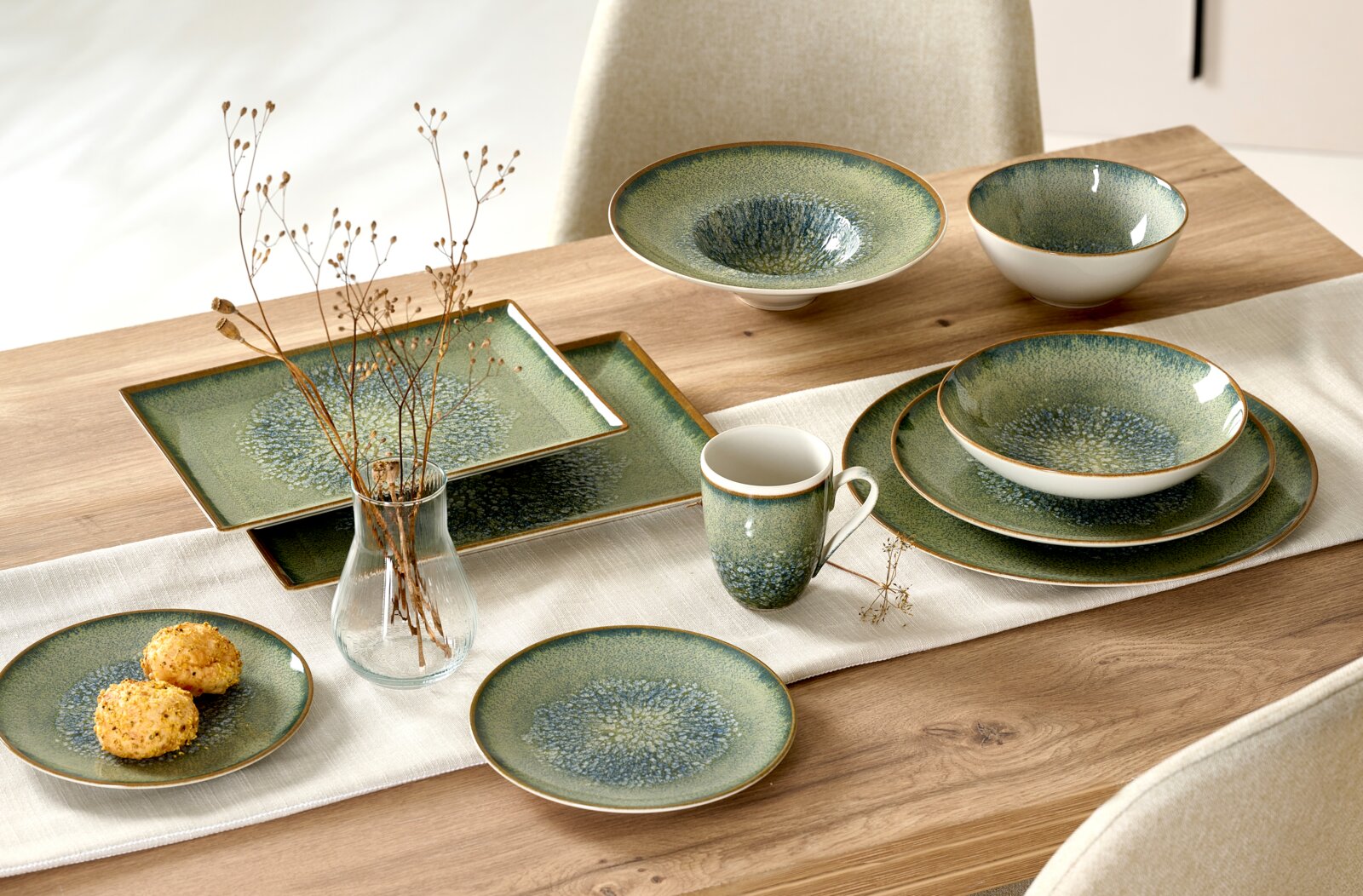 Grün-blau gesprenkeltes Keramikgeschirr mit Tellern, Schalen, Tasse und eckigen Platten, dekorativ auf einem Holztisch arrangiert, aus schräger Vogelperspektive.