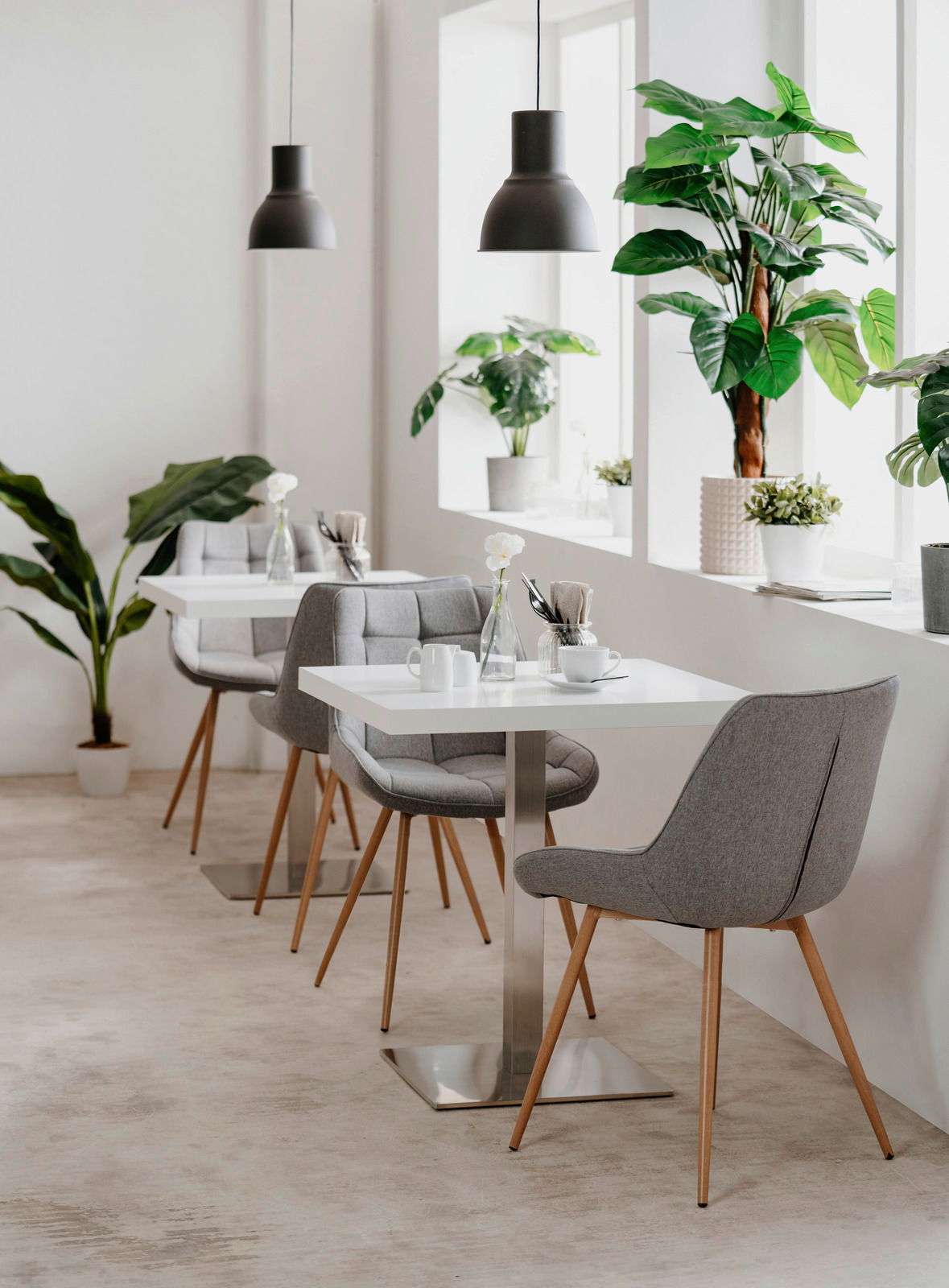 Moderner Esstisch mit weißer Tischplatte und Metallfuß, umgeben von grauen Polsterstühlen mit Holzbeinen, in einem hellen Raum mit großen Pflanzen und Hängelampen, seitliche Perspektive.