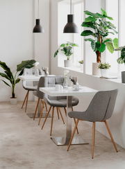 Moderner Esstisch mit weißer Tischplatte und Metallfuß, umgeben von grauen Polsterstühlen mit Holzbeinen, in einem hellen Raum mit großen Pflanzen und Hängelampen, seitliche Perspektive.
