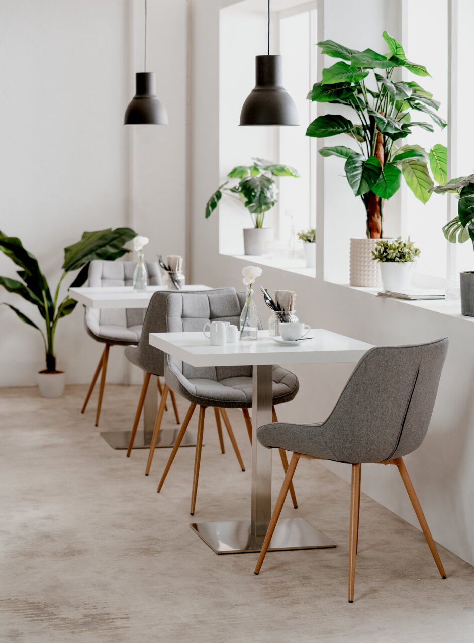 Moderner Esstisch mit weißer Tischplatte und Metallfuß, umgeben von grauen Polsterstühlen mit Holzbeinen, in einem hellen Raum mit großen Pflanzen und Hängelampen, seitliche Perspektive.
