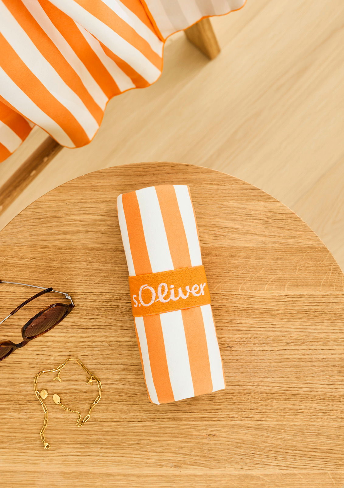 Gestreiftes Sporttuch in Orange und Weiß, zusammengerollt auf einem Holztisch, mit einer Sonnenbrille und einer goldenen Kette daneben, aus der Vogelperspektive.