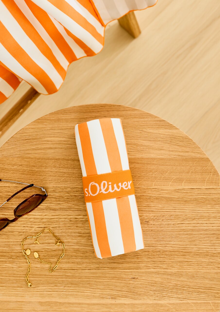 Gestreiftes Sporttuch in Orange und Weiß, zusammengerollt auf einem Holztisch, mit einer Sonnenbrille und einer goldenen Kette daneben, aus der Vogelperspektive.