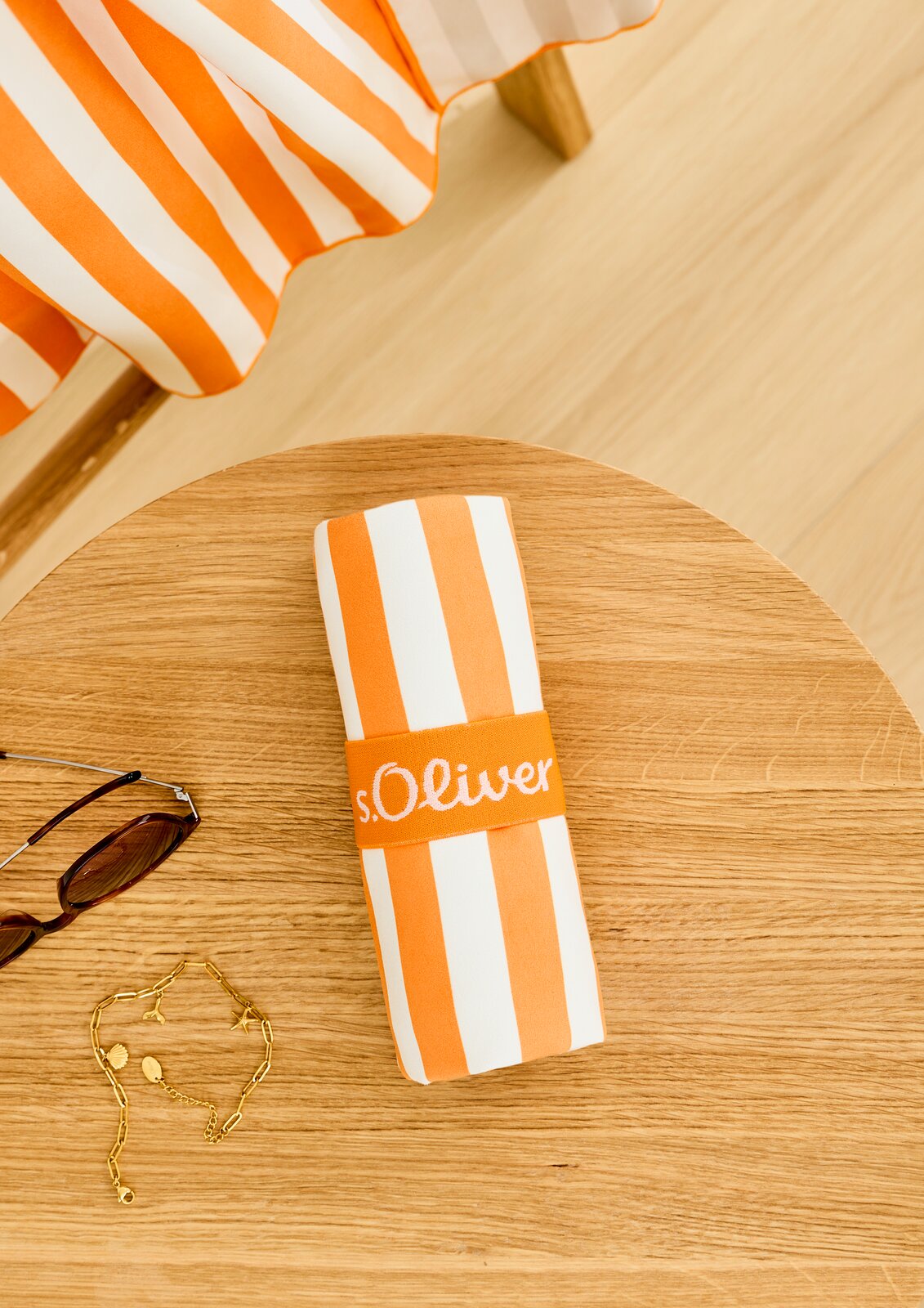 Een gestreepte sportdoek in oranje en wit, opgerold op een houten tafel, met daarnaast een zonnebril en een gouden ketting, gezien vanuit vogelperspectief.