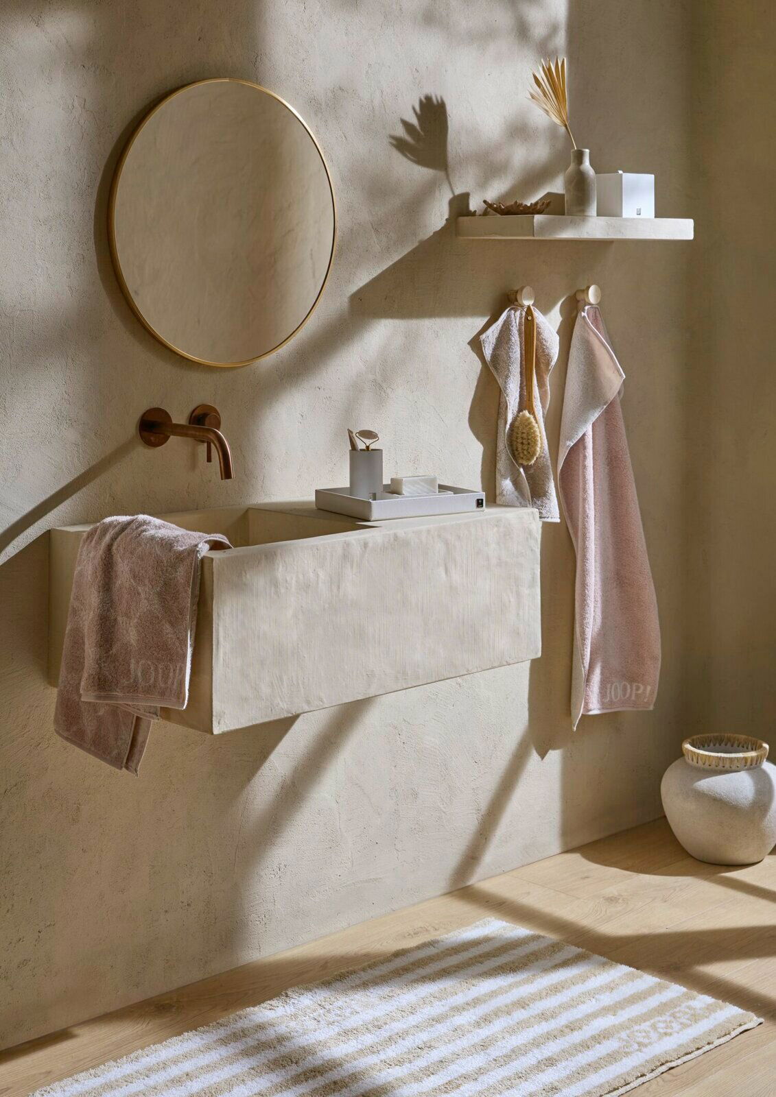 JOOP! Handtuch DOUBLEFACE Badezimmer mit beigen Handtüchern, die an Haken hängen und auf einem Waschbecken liegen, mit einem runden Spiegel und minimalistischer Dekoration. Perspektive von vorne.