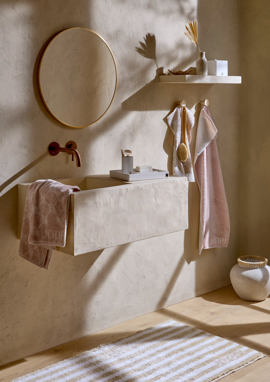 JOOP! Handtuch DOUBLEFACE Badezimmer mit beigen Handtüchern, die an Haken hängen und auf einem Waschbecken liegen, mit einem runden Spiegel und minimalistischer Dekoration. Perspektive von vorne.