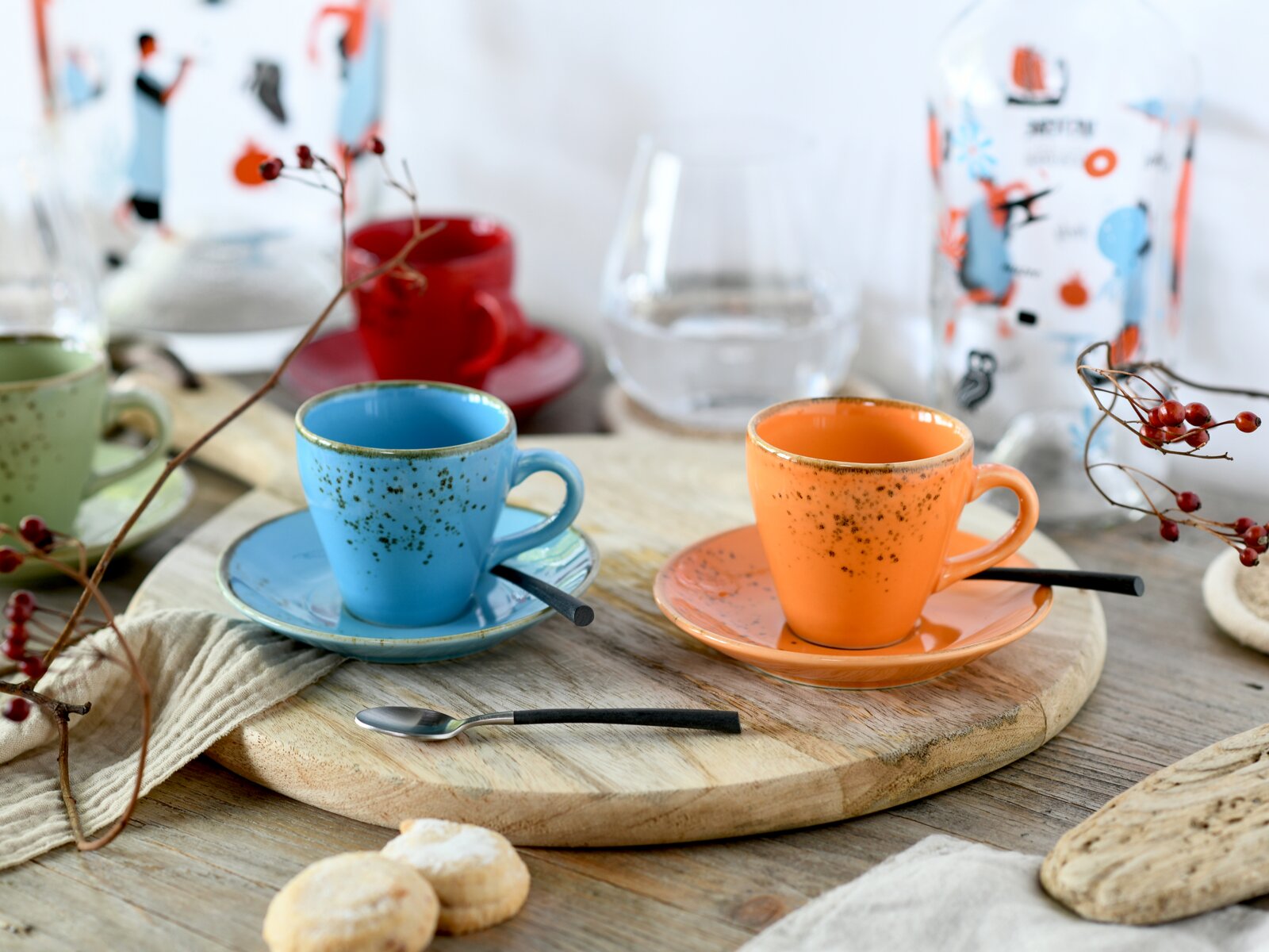 Zwei gesprenkelte Espressotassen aus Keramik in Blau und Orange mit passenden Untertassen und Löffeln auf einem runden Holzbrett, fotografiert in leicht schräger Vorderansicht auf Tischhöhe.