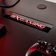 Detailaufnahme aus schräger Draufsicht: Carbonfaser-Oberfläche eines Gaming-Tisches mit rotem „DXGAMING – Designed for Players“-Logo, integrierter USB-Ladebuchse und teilweise sichtbarem Gamepad unten rechts