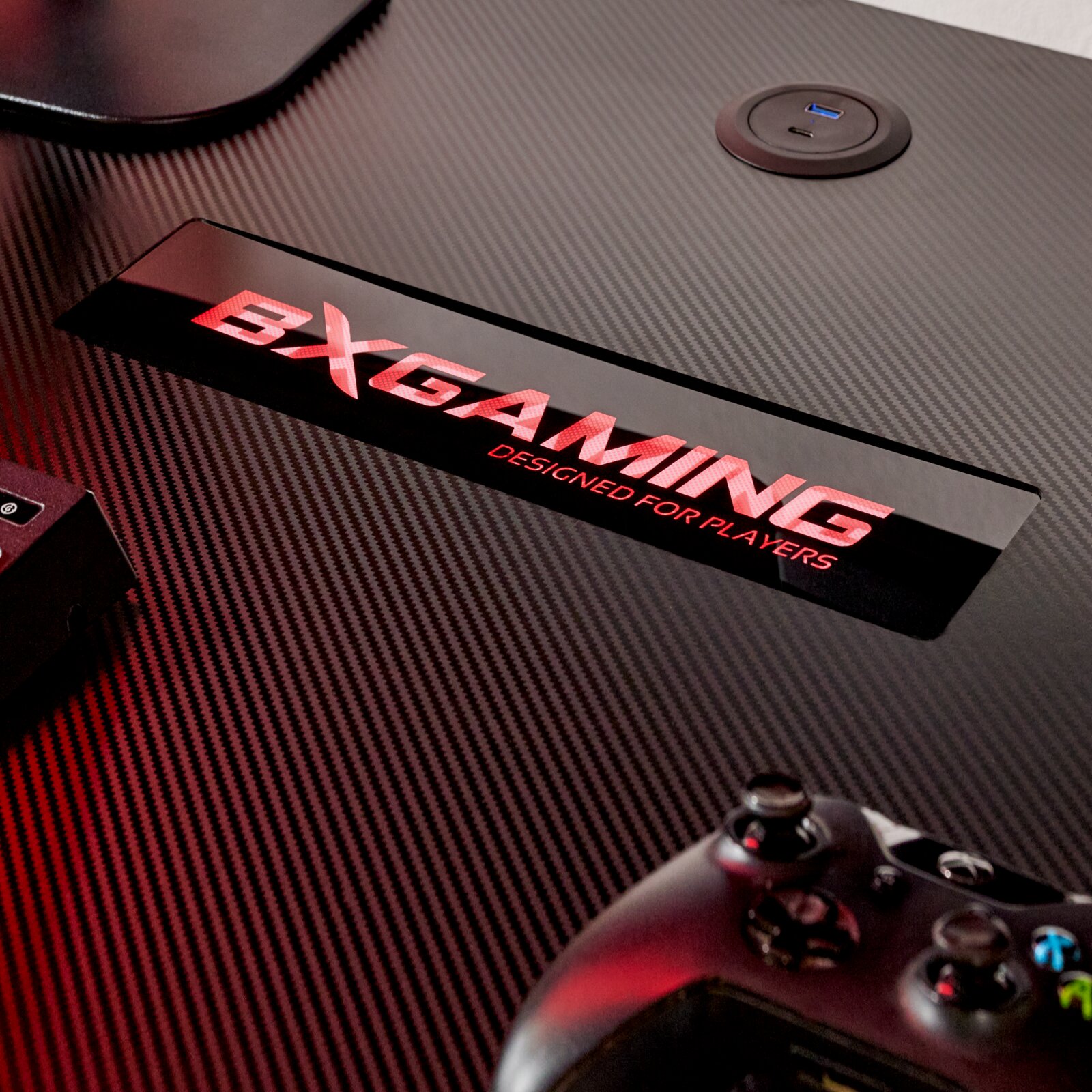 Detailaufnahme aus schräger Draufsicht: Carbonfaser-Oberfläche eines Gaming-Tisches mit rotem „DXGAMING – Designed for Players“-Logo, integrierter USB-Ladebuchse und teilweise sichtbarem Gamepad unten rechts