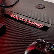Detailaufnahme aus schräger Draufsicht: Carbonfaser-Oberfläche eines Gaming-Tisches mit rotem „DXGAMING – Designed for Players“-Logo, integrierter USB-Ladebuchse und teilweise sichtbarem Gamepad unten rechts
