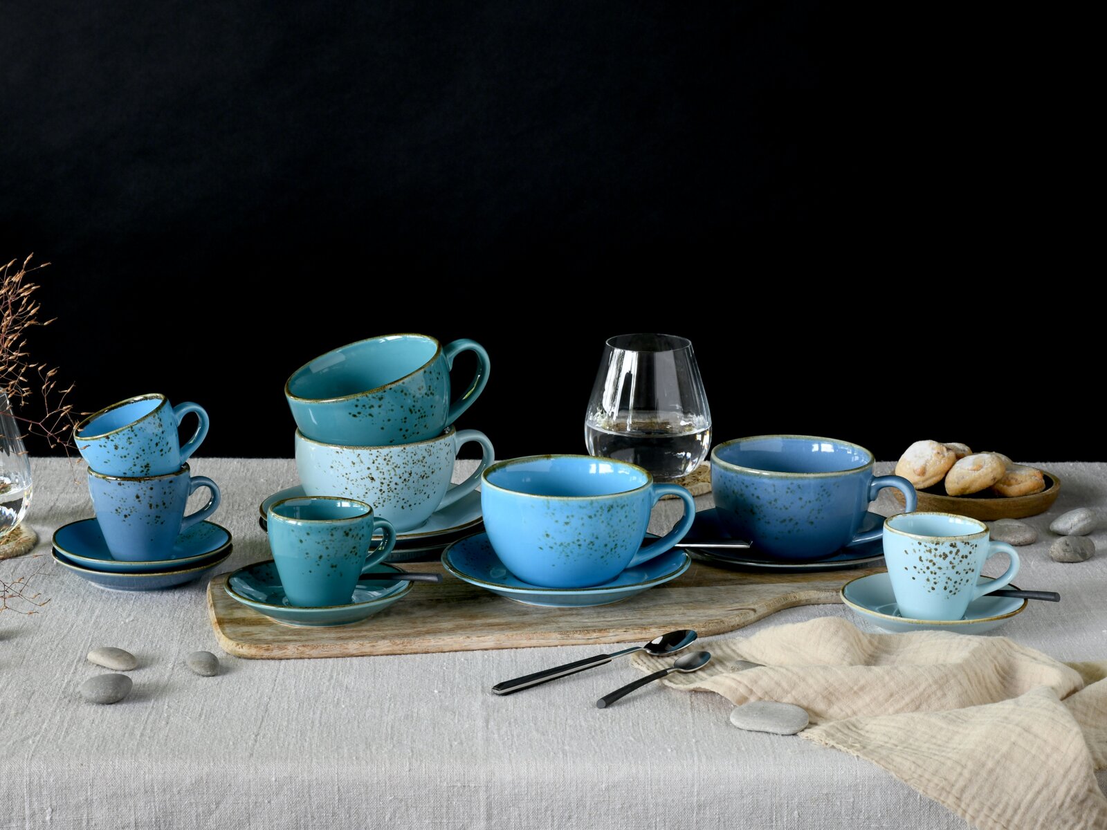 Stillleben in Frontansicht auf einem gedeckten Tisch: blaues, gesprenkeltes Keramik-Tassen- und Untertassen-Set in verschiedenen Größen, arrangiert auf einem Holzbrett vor schwarzem Hintergrund, mit Wasserglas, Löffeln und Gebäck.