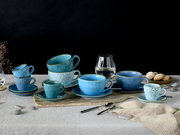 Stillleben in Frontansicht auf einem gedeckten Tisch: blaues, gesprenkeltes Keramik-Tassen- und Untertassen-Set in verschiedenen Größen, arrangiert auf einem Holzbrett vor schwarzem Hintergrund, mit Wasserglas, Löffeln und Gebäck.