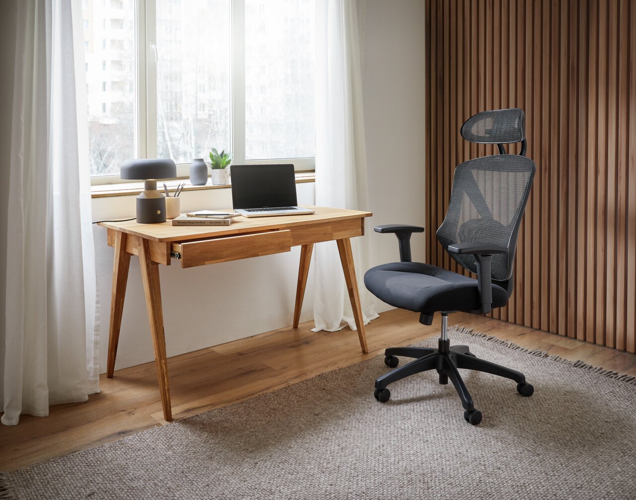 Moderner Schreibtisch aus Holz mit Schublade, Laptop und Dekorationen, daneben ein ergonomischer Bürostuhl, seitliche Perspektive.