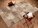 Wollteppich in natürlichen Erdtönen mit geometrischem Muster, von oben fotografiert, auf einem Holzboden mit einem kleinen Tisch und Kissen daneben.