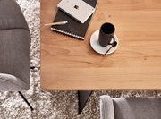 Bovenaanzicht van een lichte houten tafel in de woonkamer met een zwart koffiekopje op een wit schoteltje en een lepel, daarnaast een notitieboekje, een pen en een tablet; grijze fauteuils en een zacht tapijt op de achtergrond.