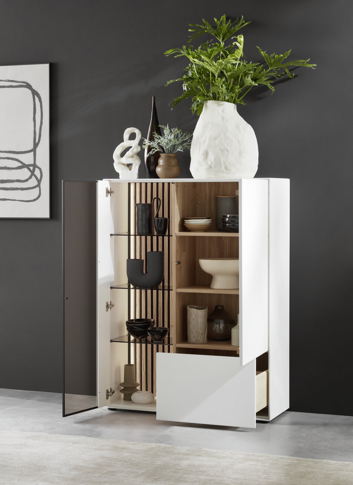 Johann Jakob Highboard (glas aan de linkerkant) LUCIANO Het moderne highboard LUCIANO van Johann Jakob met glas aan de linkerkant, gezien vanuit een open perspectief, versierd met vazen en planten, toont de planken binnenin met serviesgoed.