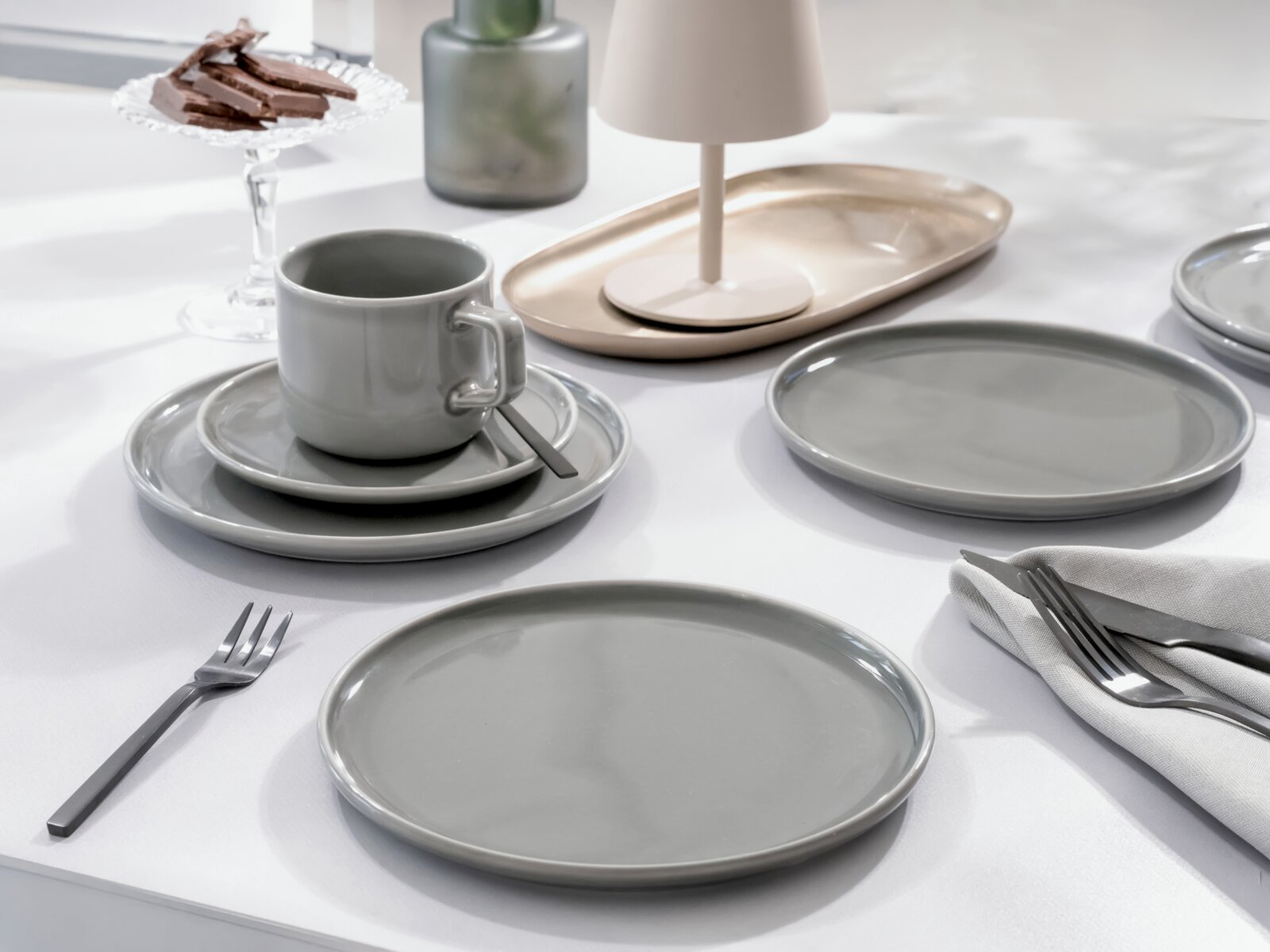 Modernes graues Keramik-Geschirrset mit Tellern, Tasse und Untertasse auf weiß gedecktem Tisch, seitliche Perspektive auf Augenhöhe mit leicht schrägem Blick von vorn.