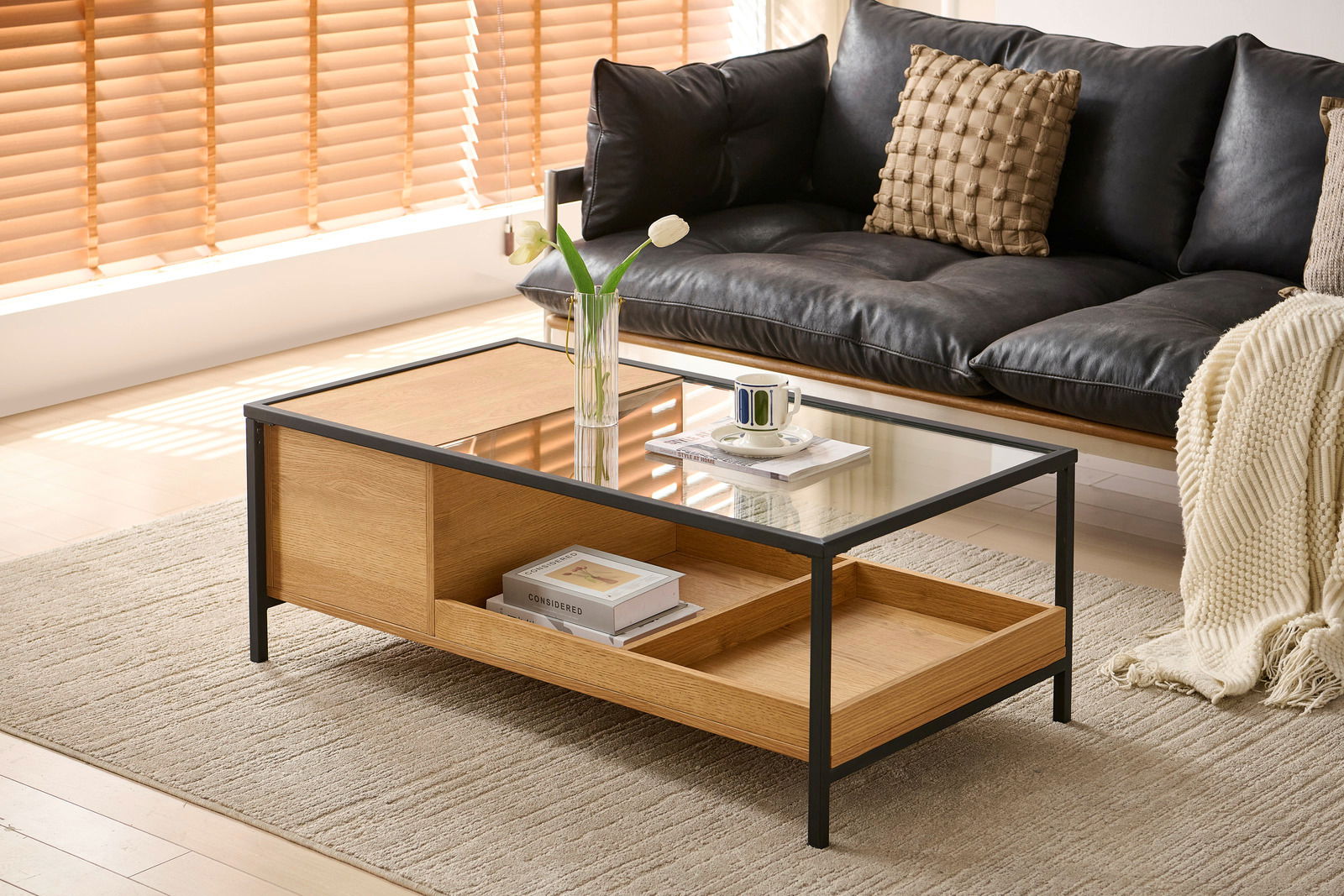 Moderner Couchtisch aus Holz und Glas mit schwarzem Metallrahmen, seitliche Perspektive, dekoriert mit einer Vase und einer Tasse auf einem Teppich vor einem Sofa.