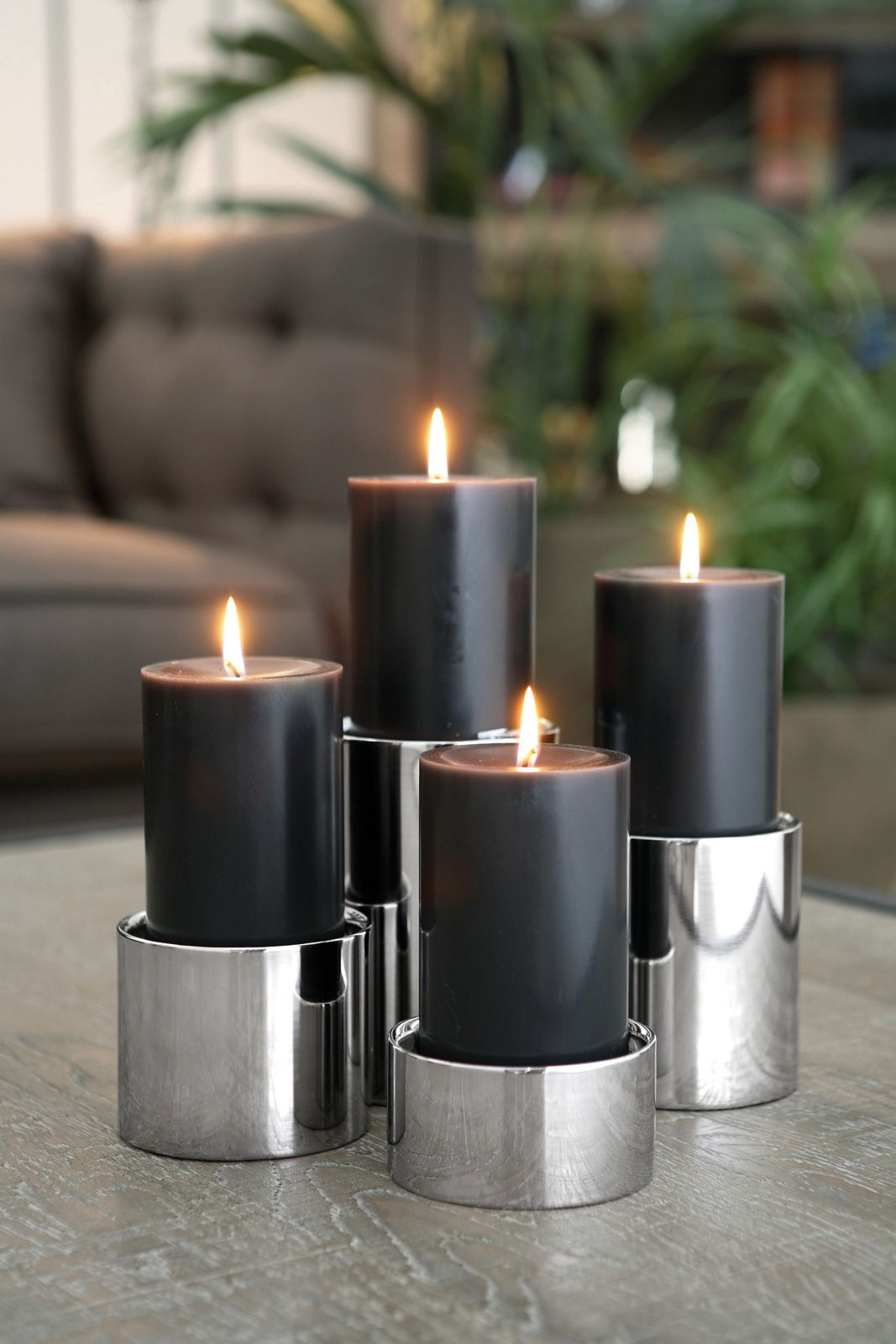 Vier schwarze Kerzen in Edelstahl-Stumpenhaltern, auf einem Tisch arrangiert, mit unscharfem Hintergrund. Perspektive: seitlich.