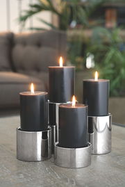 Vier schwarze Kerzen in Edelstahl-Stumpenhaltern, auf einem Tisch arrangiert, mit unscharfem Hintergrund. Perspektive: seitlich.
