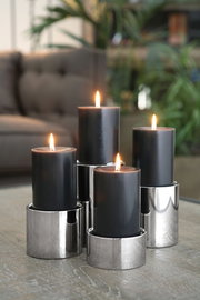 Vier schwarze Kerzen in Edelstahl-Stumpenhaltern, auf einem Tisch arrangiert, mit unscharfem Hintergrund. Perspektive: seitlich.