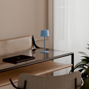 Blaue, minimalistische Tischlampe auf Glas-Schreibtisch neben geschlossenem Laptop, Innenraum mit Lederbank und Fenster im Hintergrund, seitliche Perspektive