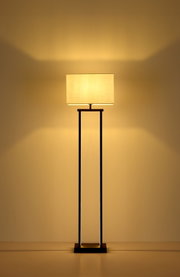 Moderne Stehleuchte mit rechteckigem, cremefarbenem Stoffschirm und schlankem schwarzem Metallgestell, warm leuchtend vor beiger Wand, frontal aus Augenhöhe fotografiert.