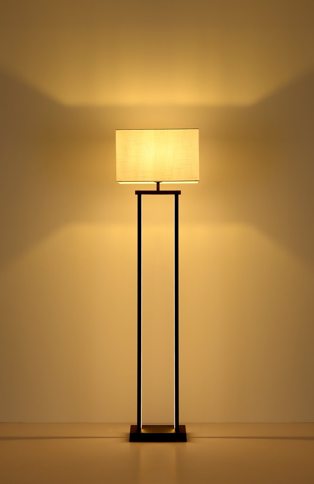 Moderne Stehleuchte mit rechteckigem, cremefarbenem Stoffschirm und schlankem schwarzem Metallgestell, warm leuchtend vor beiger Wand, frontal aus Augenhöhe fotografiert.