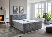 Graues Boxspringbett BX2200 CHESTER in einem modernen Schlafzimmer mit Kamin und großen Fenstern, seitliche Perspektive.