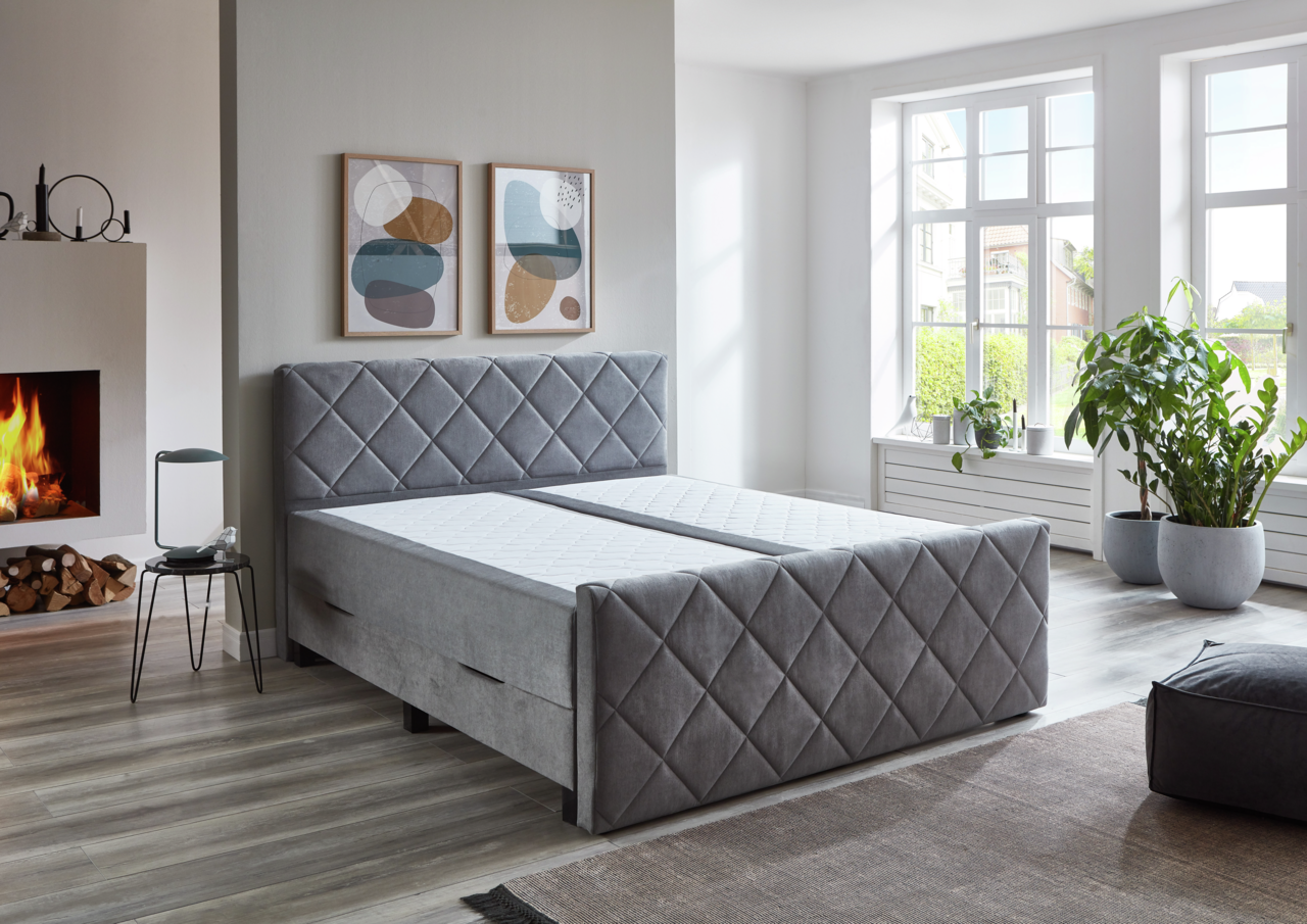 Graues Boxspringbett BX2200 CHESTER in einem modernen Schlafzimmer mit Kamin und großen Fenstern, seitliche Perspektive.