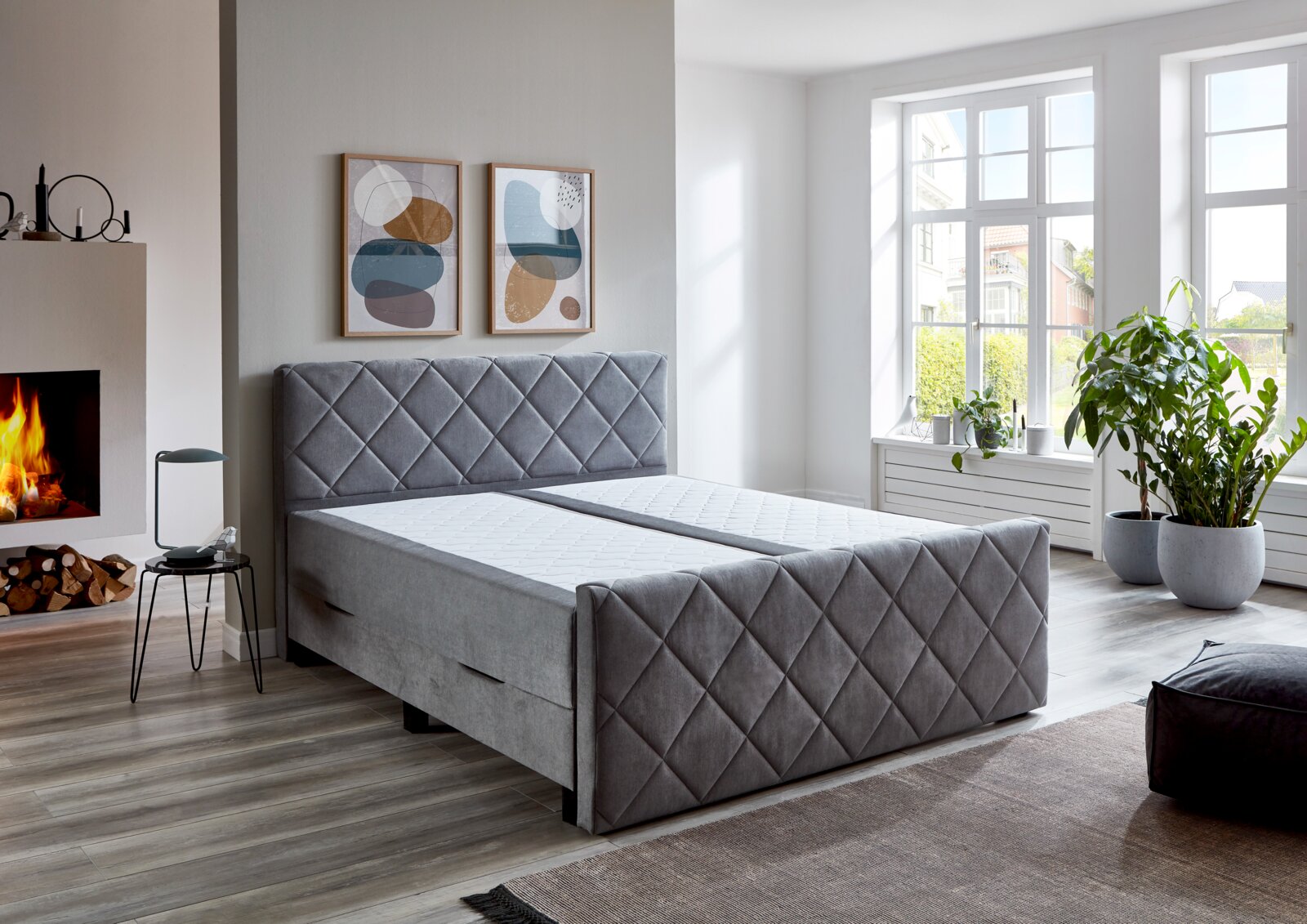 Graues Boxspringbett BX2200 CHESTER in einem modernen Schlafzimmer mit Kamin und großen Fenstern, seitliche Perspektive.