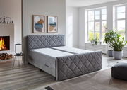 Graues Boxspringbett BX2200 CHESTER in einem modernen Schlafzimmer mit Kamin und großen Fenstern, seitliche Perspektive.
