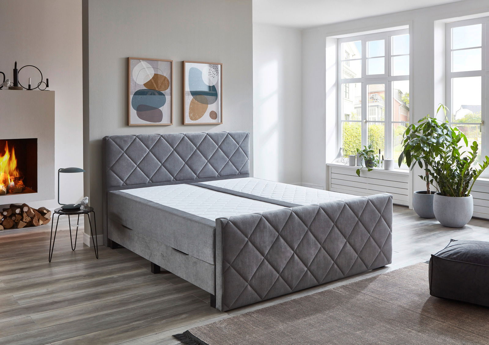 Graues Boxspringbett BX2200 CHESTER in einem modernen Schlafzimmer mit Kamin und großen Fenstern, seitliche Perspektive.
