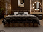 Een luxe gestoffeerd bedframe in een stijlvol ingerichte slaapkamer, gefotografeerd van voren. Het bed is voorzien van een donkere, gestoffeerde deken en verschillende kussens. Op de achtergrond zijn een verlichte muur, een ronde spiegel en een nachtkastj