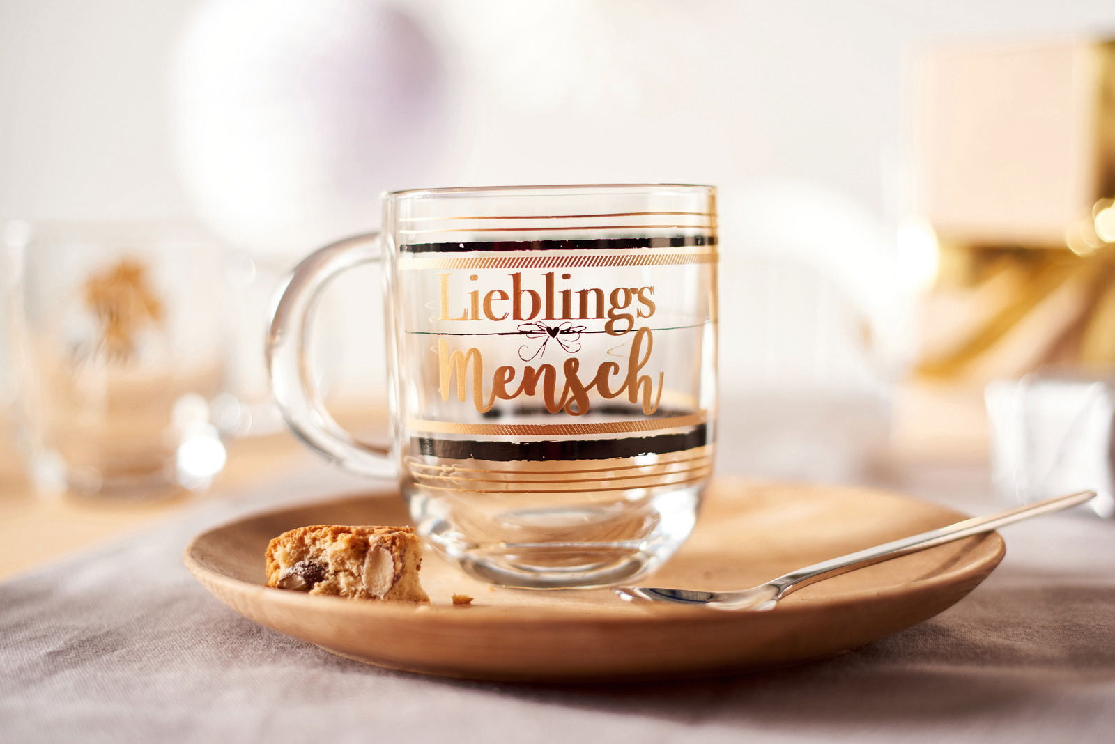 Glas-Tasse mit der Aufschrift 'Lieblingsmensch' in goldener Schrift, auf einem Holzteller mit einem Keks und einem Löffel, aus seitlicher Perspektive fotografiert.