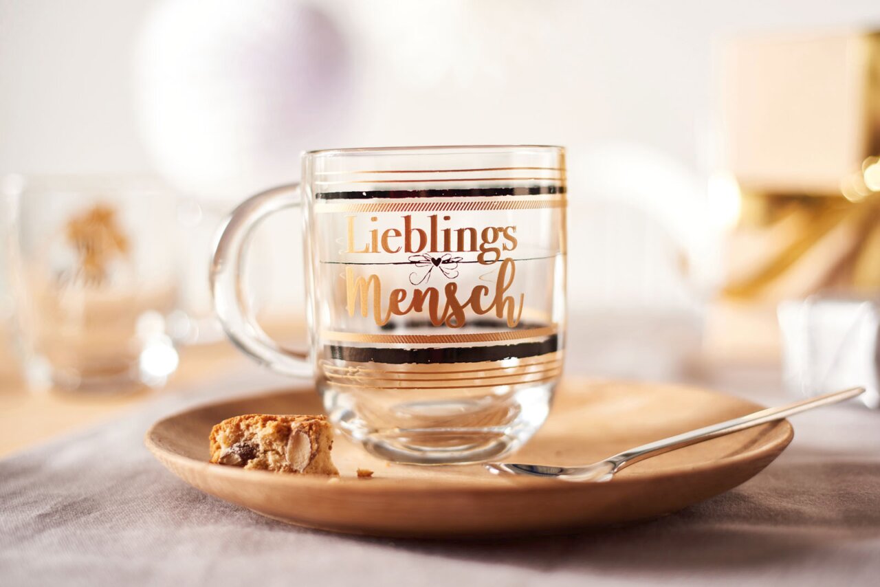 Glas-Tasse mit der Aufschrift 'Lieblingsmensch' in goldener Schrift, auf einem Holzteller mit einem Keks und einem Löffel, aus seitlicher Perspektive fotografiert.