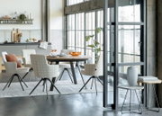Heller Essbereich im Industrial-Loft mit rundem Holzesstisch und gepolsterten Stühlen in Creme- und Musterstoff, aufgenommen in leichter Schrägansicht von vorne rechts.