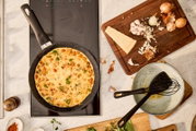 Bratpfanne mit Omelett auf einem Herd von oben betrachtet, umgeben von Zutaten wie Zwiebeln, Knoblauch und Parmesan auf einem Schneidebrett.