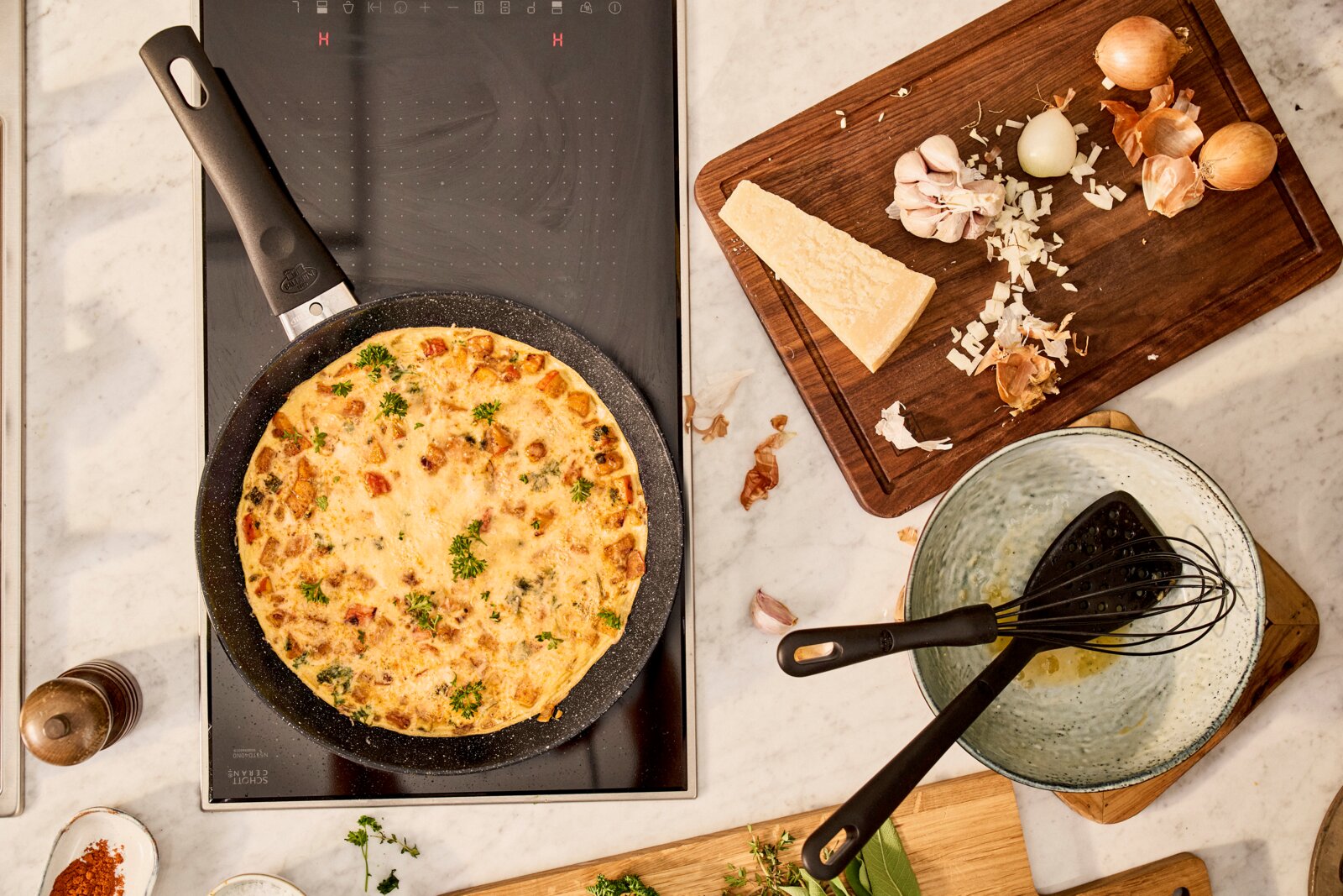 Bratpfanne mit Omelett auf einem Herd von oben betrachtet, umgeben von Zutaten wie Zwiebeln, Knoblauch und Parmesan auf einem Schneidebrett.