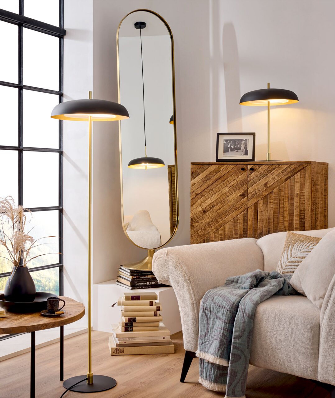 Moderne staande lamp in zwart en goud met een platte kap, warm licht en een ronde voet, naast de bank en het raam; perspectief: licht zijdelings vooraanzicht in de woonkamer.