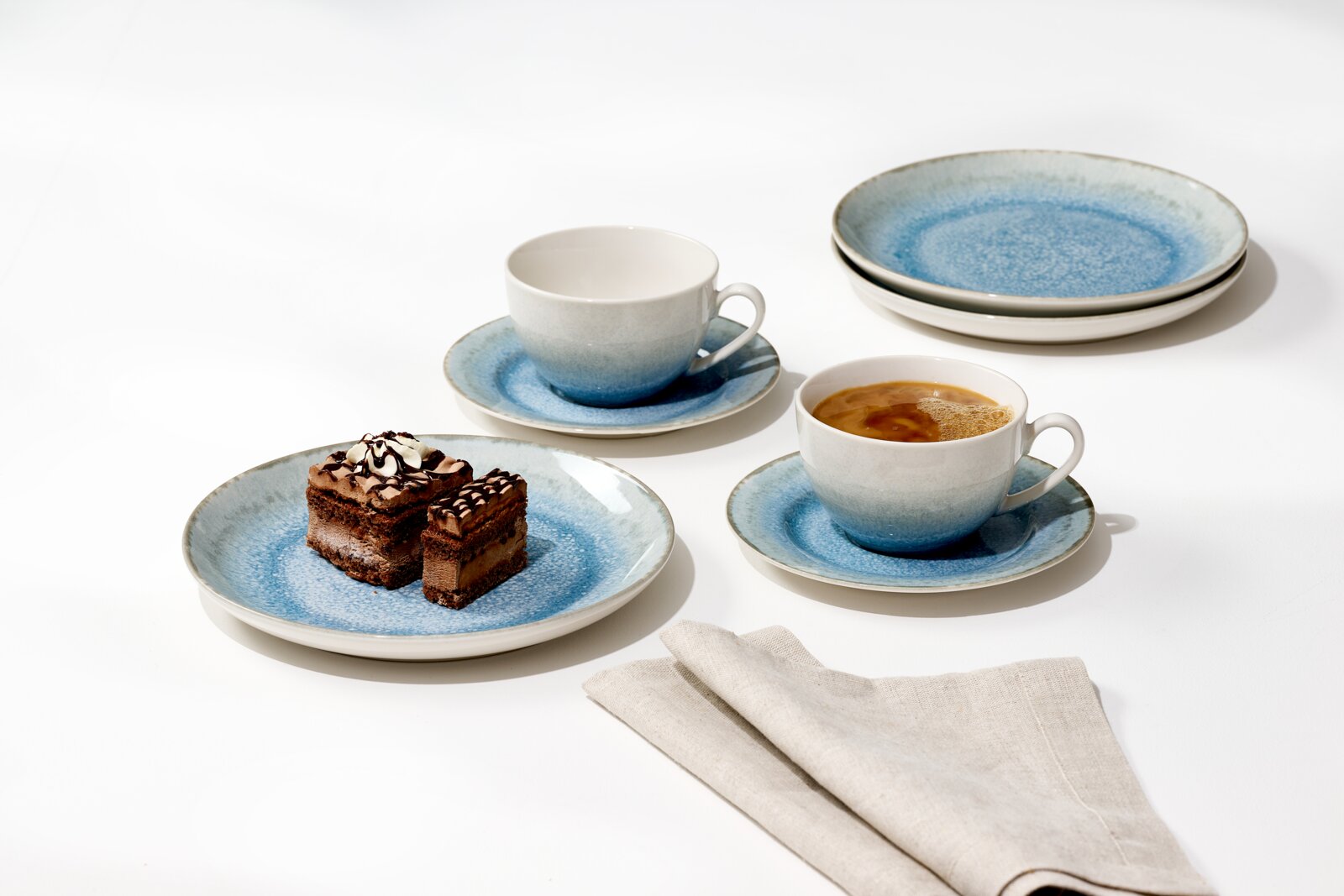Studioaufnahme in leicht erhöhter Schrägansicht: blau-weißes Keramik-Geschirrset mit zwei Tassen auf Untertassen, zwei Tellern und Dessertteller mit Schokokuchen auf weißem Hintergrund.