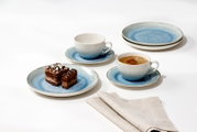 Studioaufnahme in leicht erhöhter Schrägansicht: blau-weißes Keramik-Geschirrset mit zwei Tassen auf Untertassen, zwei Tellern und Dessertteller mit Schokokuchen auf weißem Hintergrund.