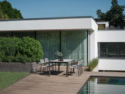 Moderner Stapelsessel auf einer Terrasse neben einem Tisch, mit Blick auf einen Garten und ein modernes Haus im Hintergrund.