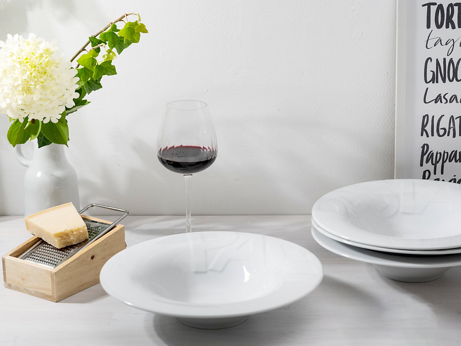 Weiße, tiefe Porzellan-Suppenteller mit dezentem Reliefmuster auf Tisch, daneben Rotweinglas, Käse und Blumenvase; Perspektive: leicht erhöhte Frontansicht.