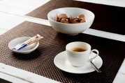 Weiße Porzellantasse mit Kaffee auf Untertasse samt Löffel, dazu eine Schale mit Schokoladenpralinen und ein kleines Ablageschälchen, aus schräger Vorderansicht von oben auf braunen Tischsets präsentiert