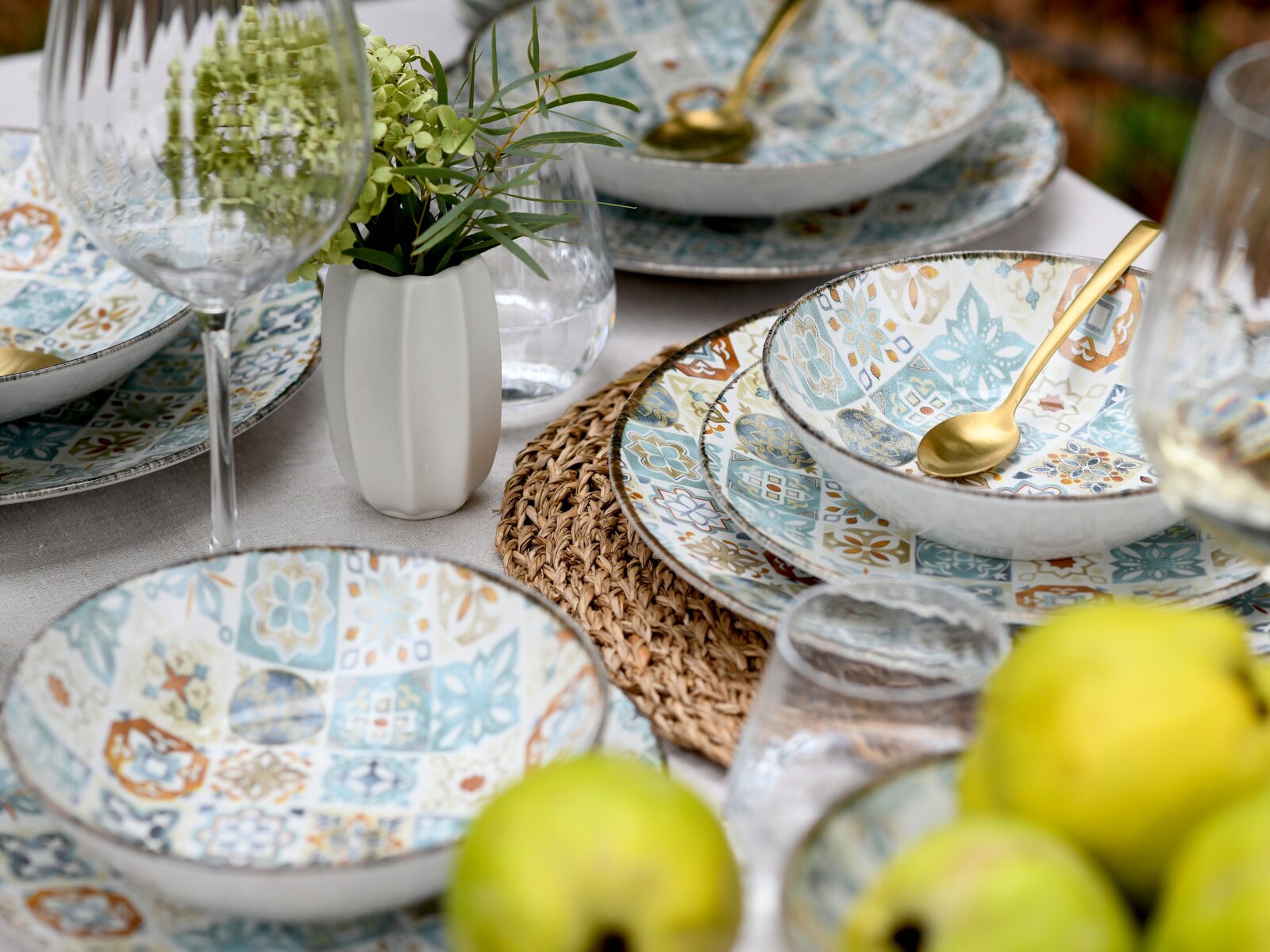 Schräg von oben fotografierte Tischszene mit gemustertem Geschirr-Set (Schalen und Teller im Patchwork-Design in Blau/Beige), goldfarbenen Löffeln, Gläsern, geflochtenem Platzset und kleiner Vase mit grünen Blumen.
