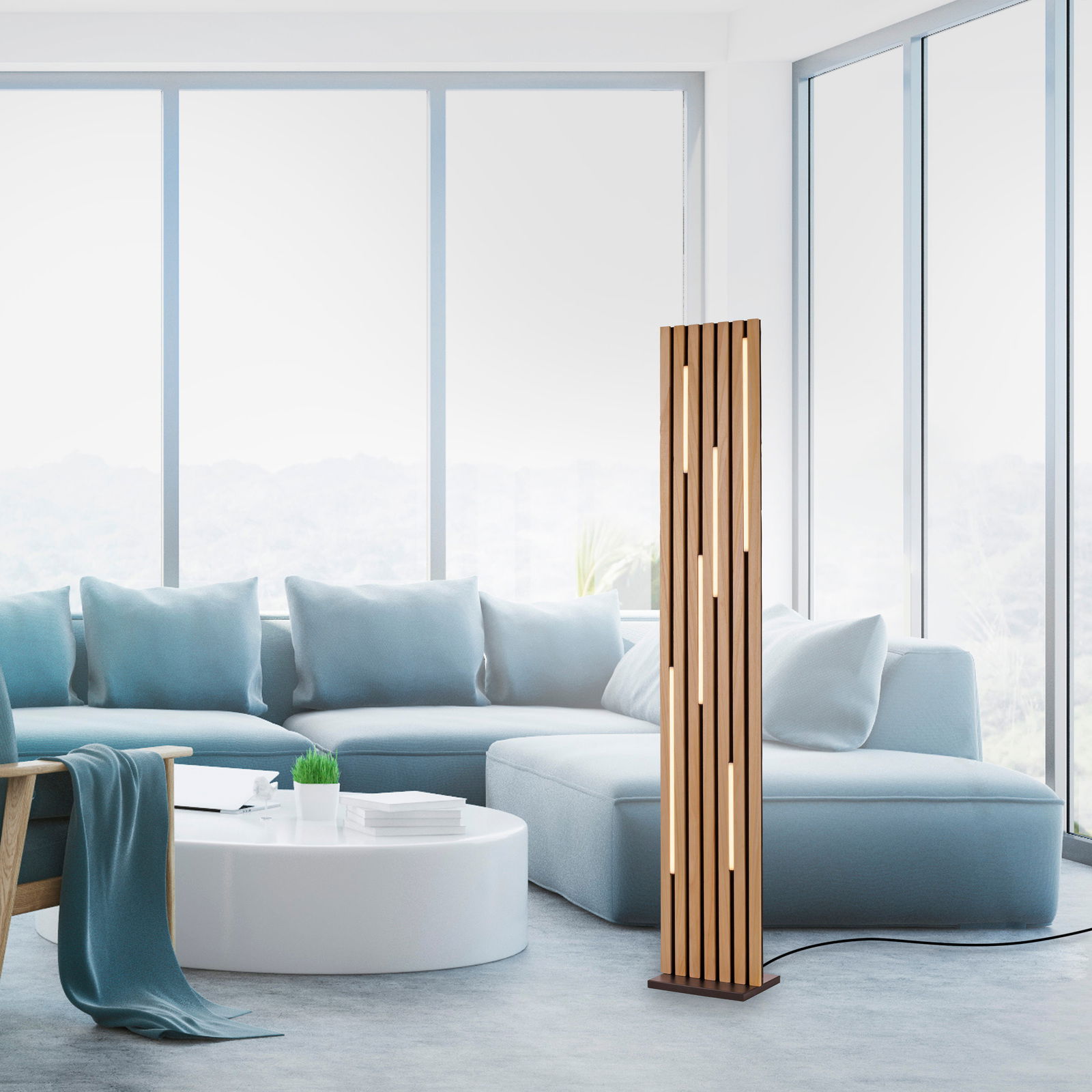 Moderne LED-Stehleuchte aus Holz mit vertikalen Schlitzen, in einem hellen Wohnzimmer mit großen Fenstern und blauen Sofas, seitliche Perspektive.