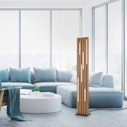 Moderne LED-Stehleuchte aus Holz mit vertikalen Schlitzen, in einem hellen Wohnzimmer mit großen Fenstern und blauen Sofas, seitliche Perspektive.