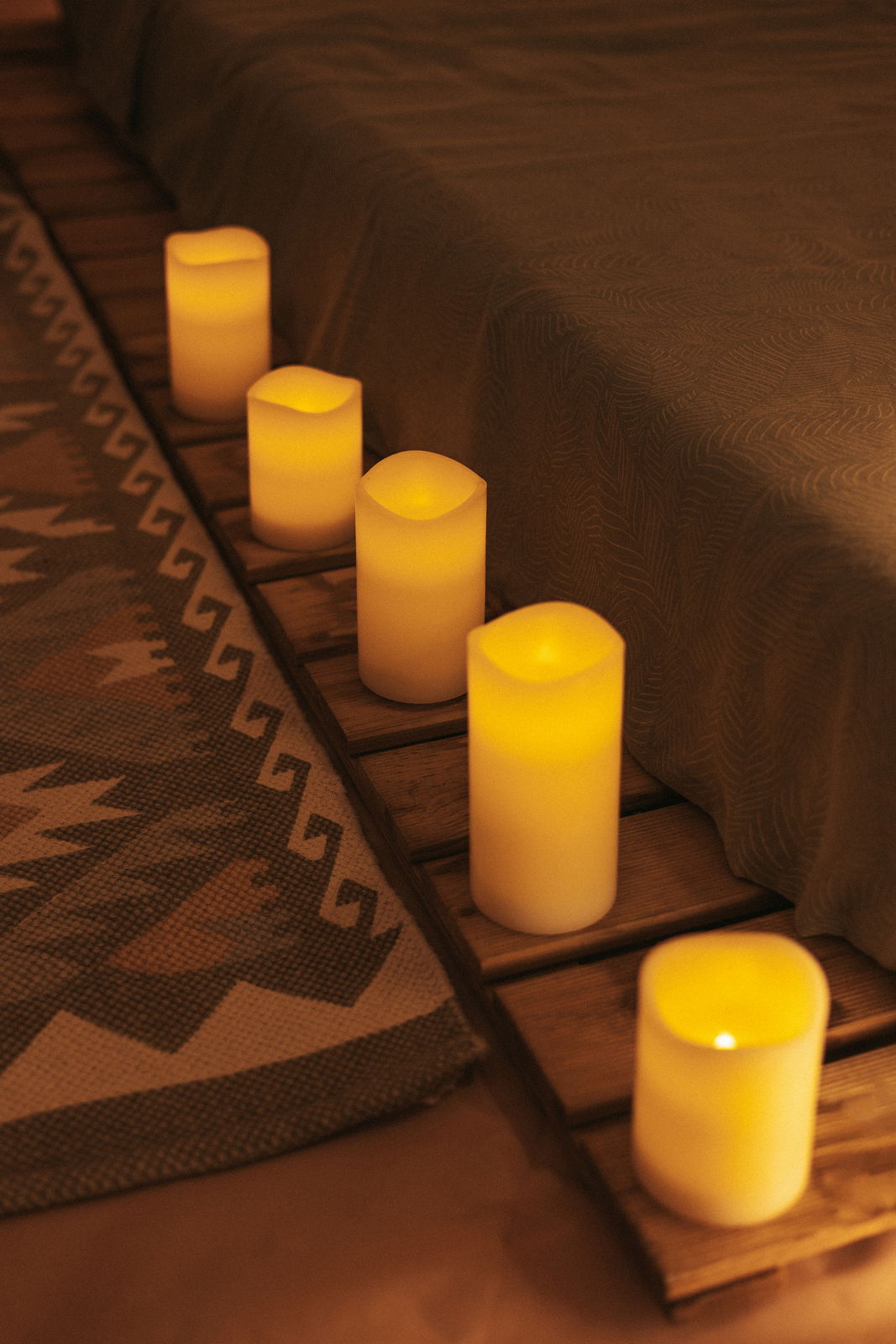 Sechs leuchtende Outdoor-Kerzen in einer gemütlichen Abendatmosphäre, auf einem Holzbrett neben einem gemusterten Teppich arrangiert, seitliche Perspektive.